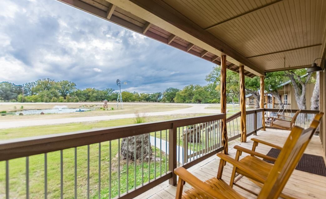 Front porch with wooden brown railing, wooden rocking chairs, porch swing, and view of small pond with fountain, windmill, and trees in the background